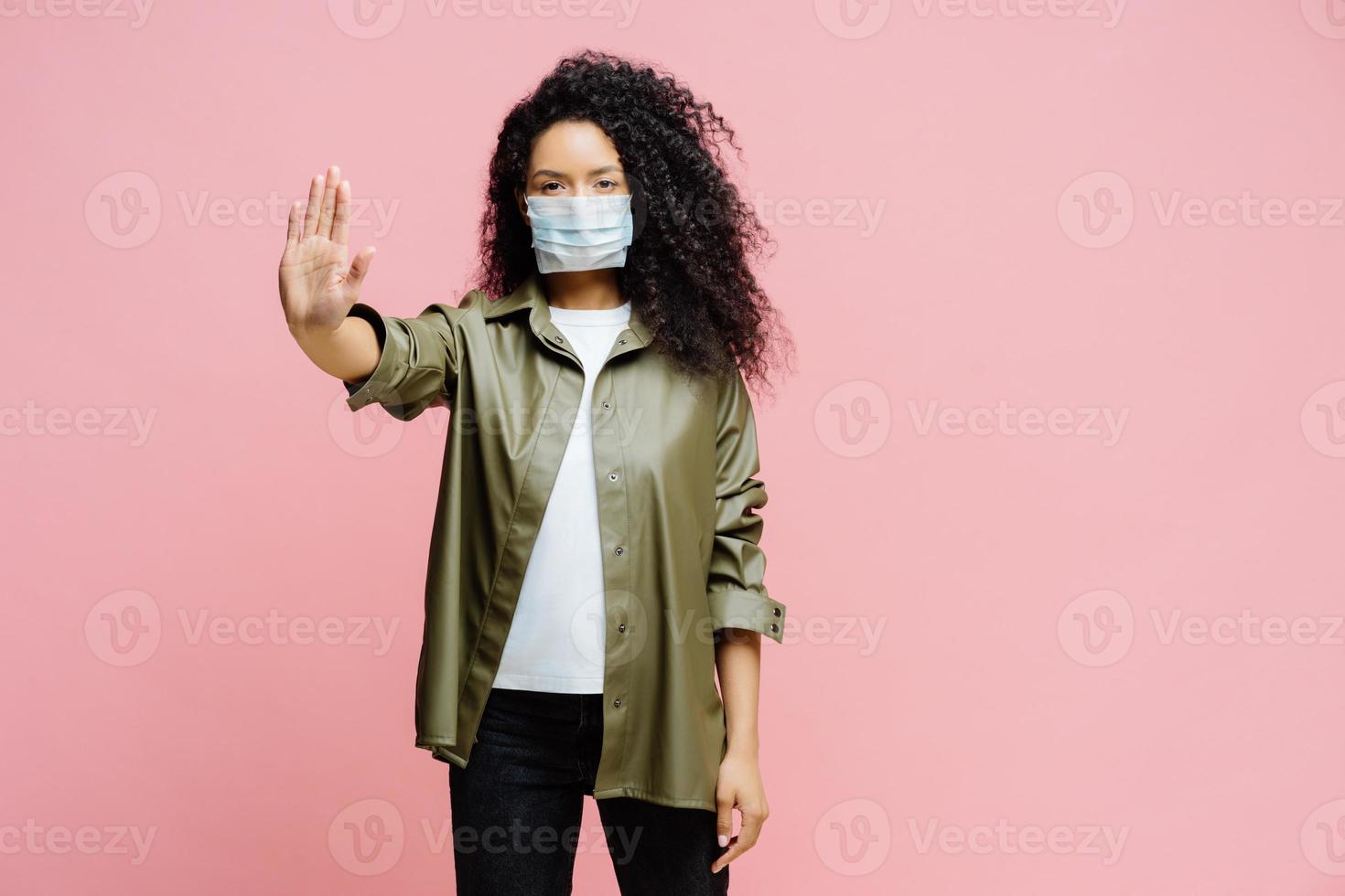 tiro horizontal de mulher afro-americana mantém a palma da mão em direção à câmera, faz gesto de parada, tenta prevenir coronavírus ou covid-19, usa máscara estéril protetora, diz não à doença pandêmica mundial foto