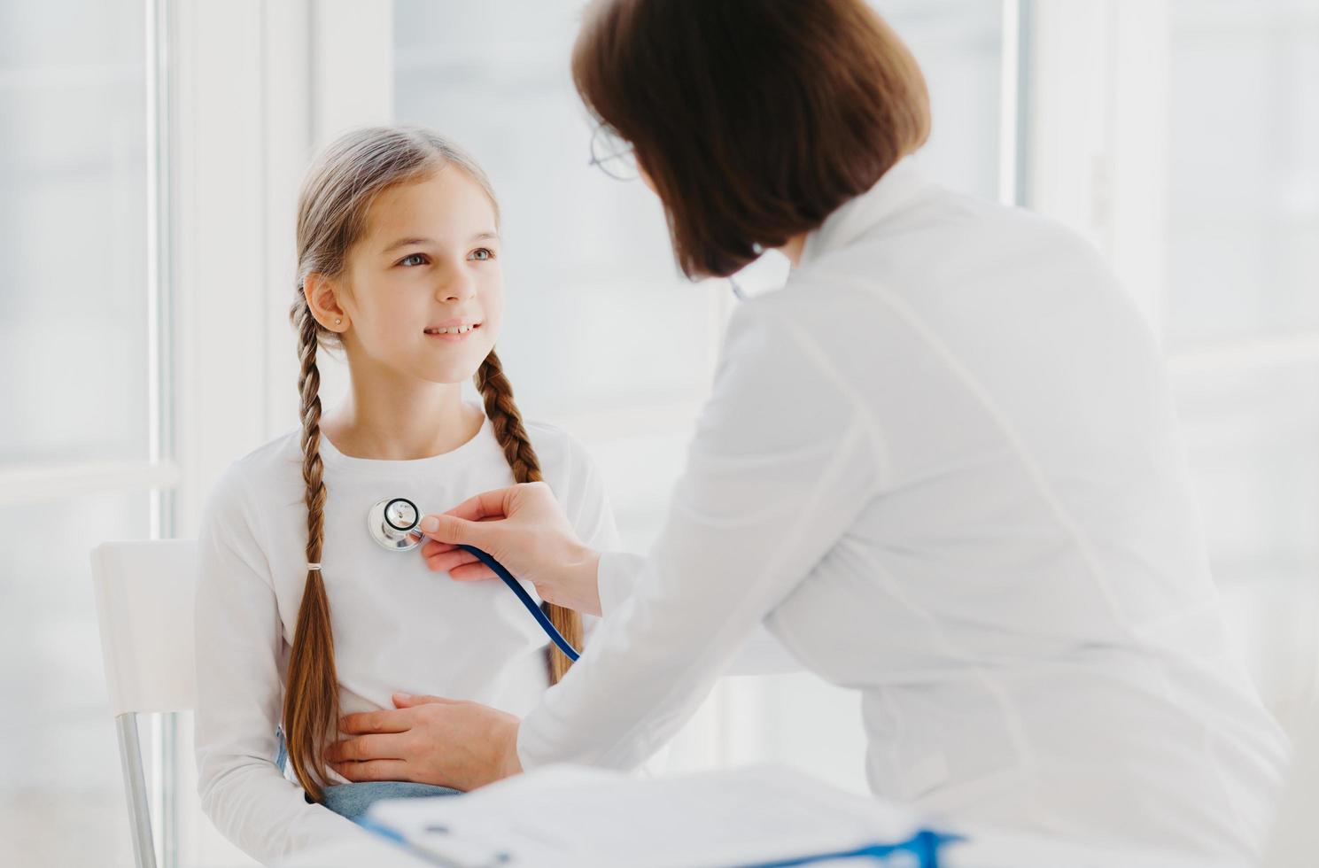 o pediatra feminino atencioso ouve os pulmões ou o coração da criança com estetoscópio. menina vem ao exame em clínica privada, tem visita médica todos os anos, explica os sintomas de sua doença foto