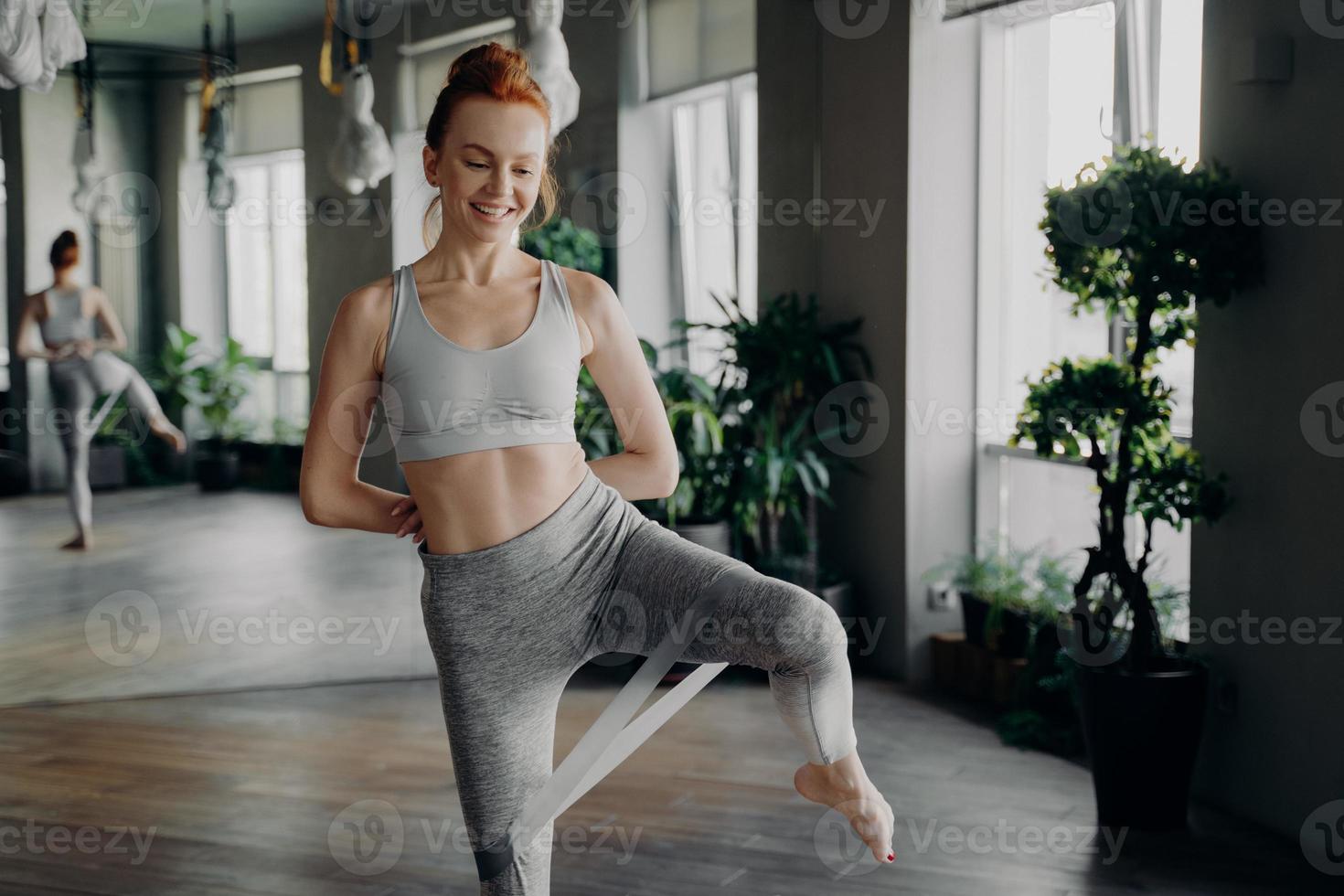 mulher magra bonita fitness com cabelo vermelho fazendo exercício de perna com elástico de resistência foto