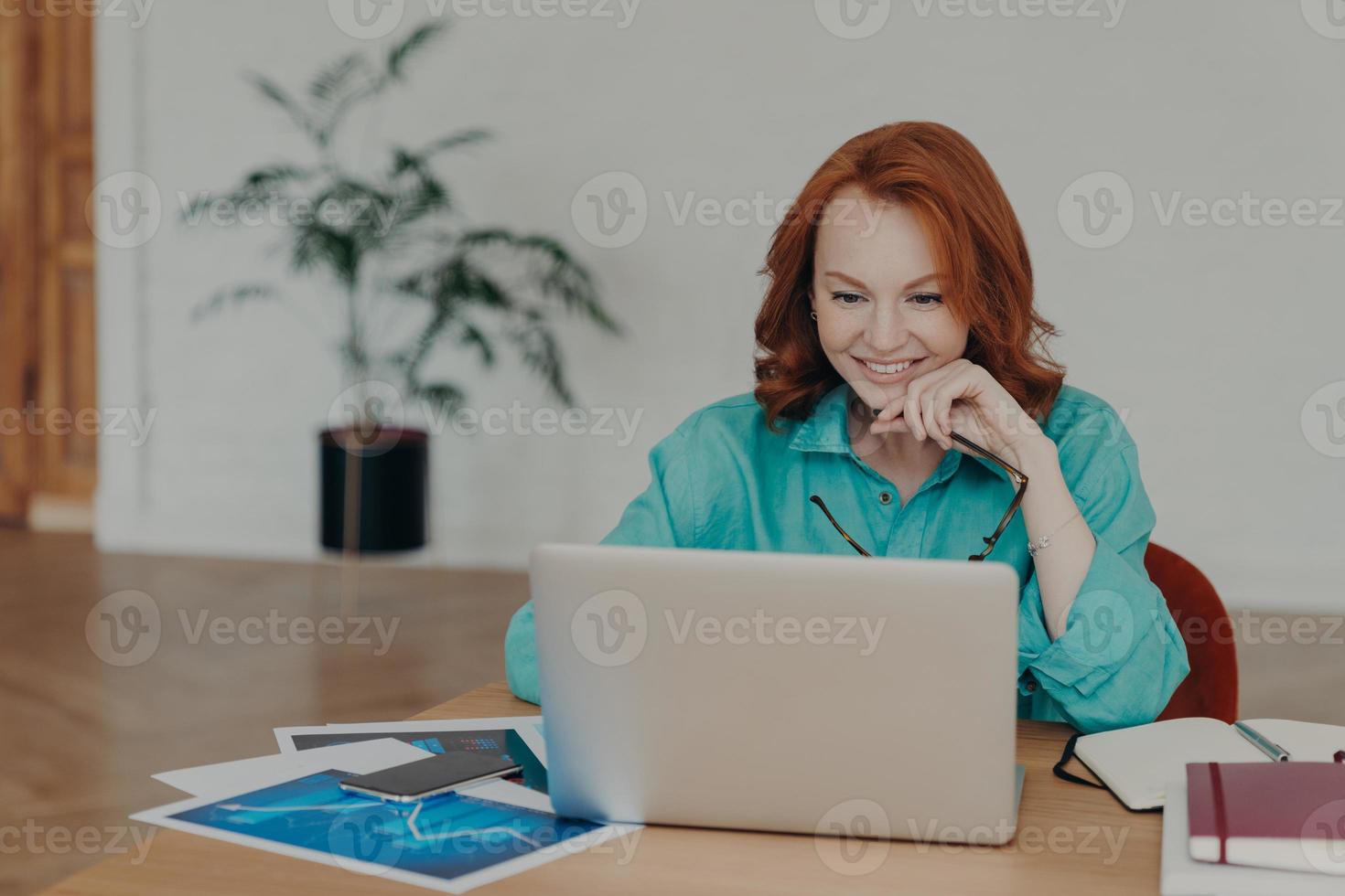 mulher milenar sorridente com cabelo ruivo trabalha ou estuda em um laptop moderno, navega na internet, tem videoconferência, dá consultoria on-line a clientes, recebe promoção de emprego ou boa oferta foto