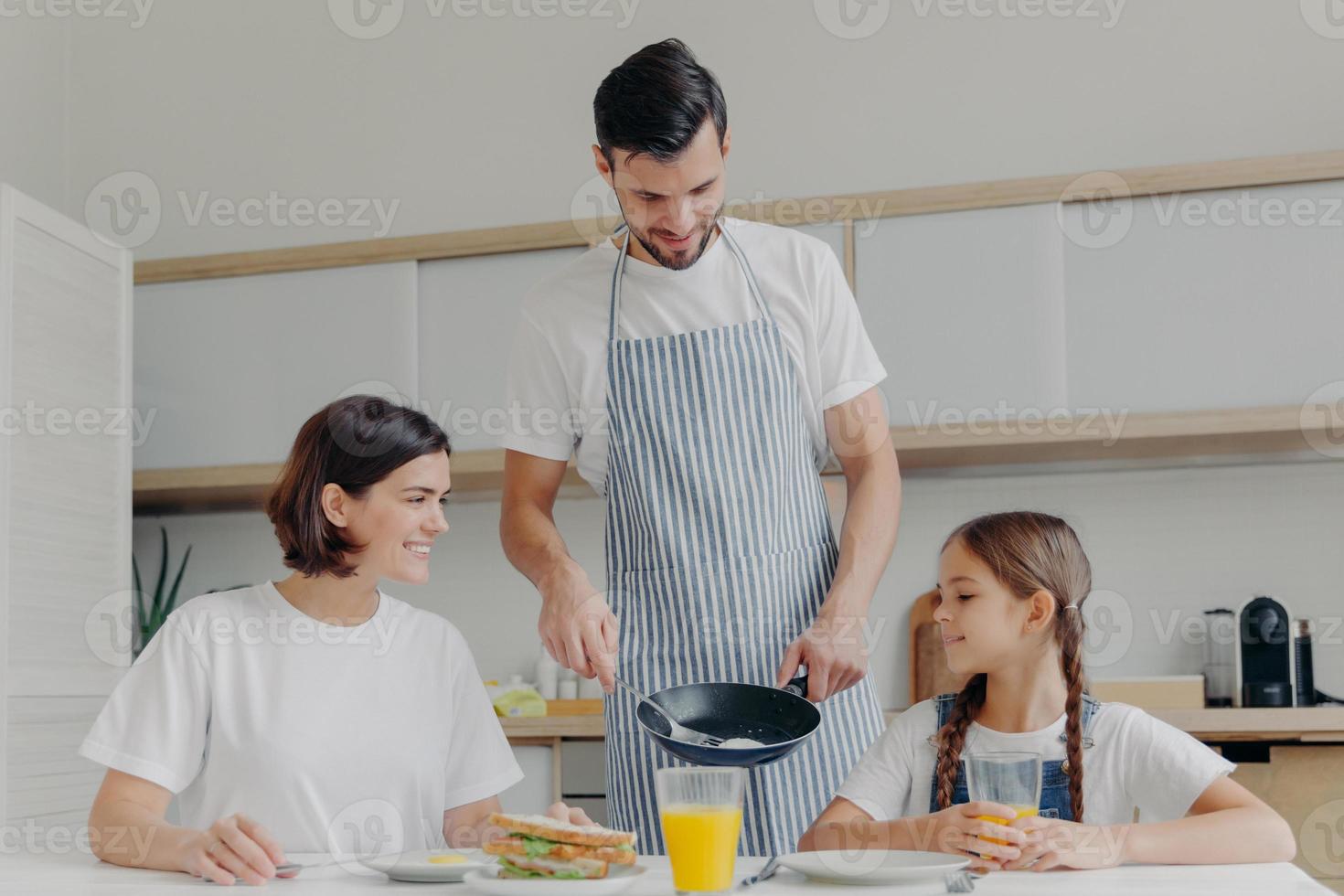 ocupado pai carinhoso prepara delicioso café da manhã para esposa e filha, usa avental, coloca ovos fritos no prato. alegre mãe e filho conversam enquanto se sentam à mesa da cozinha, fazem a refeição foto