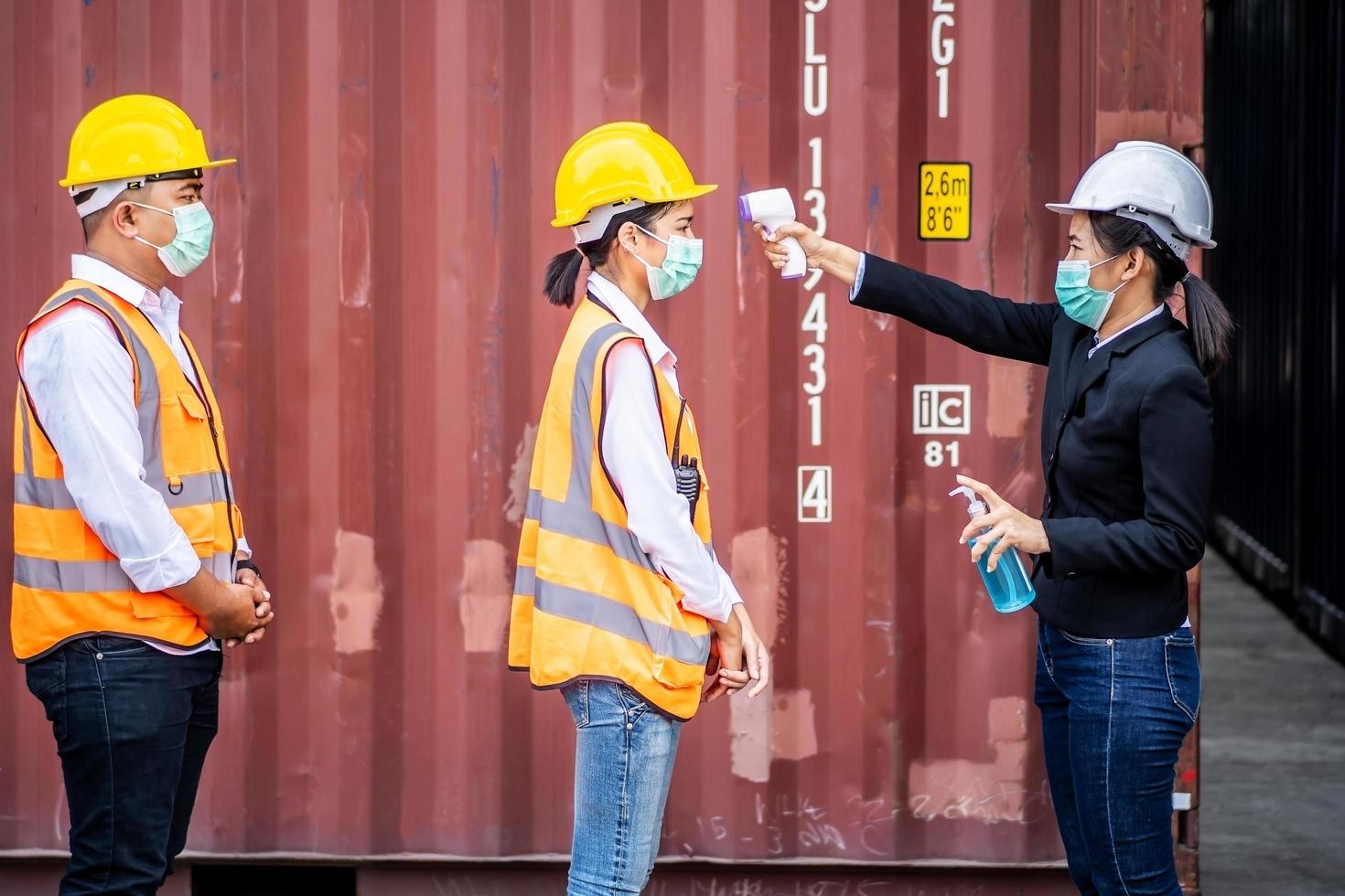 capataz feminina usando uma máscara mede a temperatura para um trabalhador em traje de segurança. eles se levantaram e esperaram a medição com um termômetro infravermelho sem contato para evitar o vírus covid foto