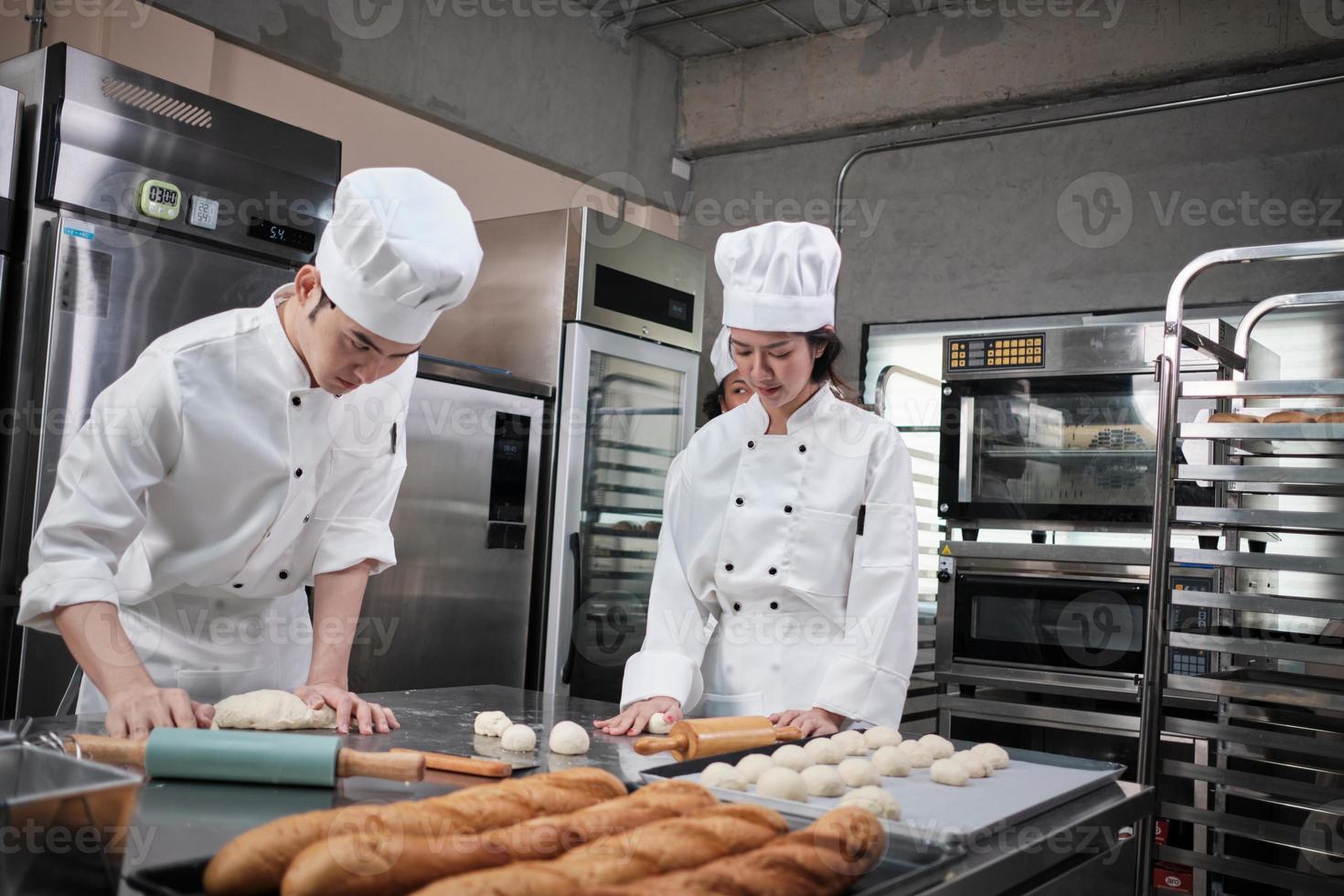 dois chefs asiáticos profissionais em uniformes e aventais de cozinheiro branco amassam massa e ovos, preparam pão, biscoitos e alimentos frescos de padaria, assando em um forno em uma cozinha de aço inoxidável de restaurante. foto