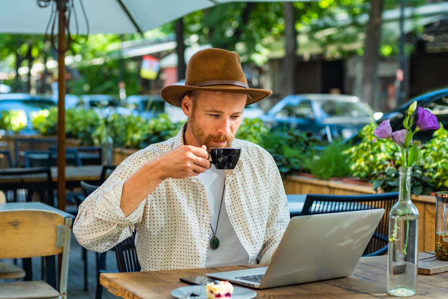 jovem hipster elegante no trabalho de chapéu com laptop ao ar livre no café de rua foto