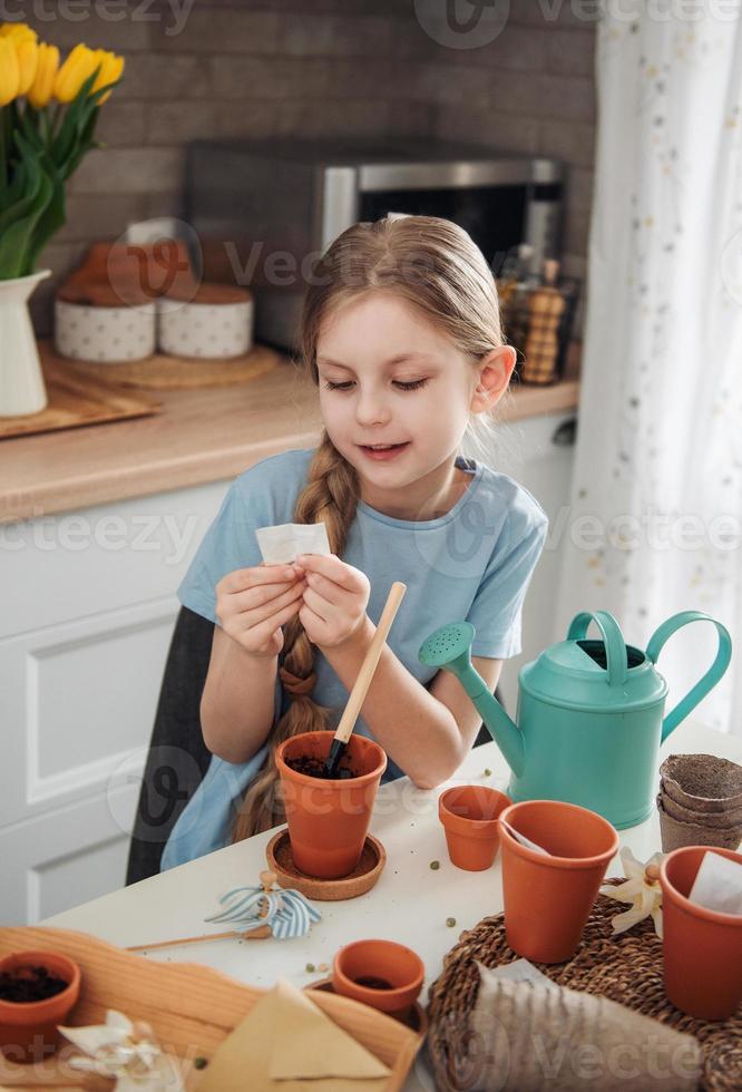 menina sentada à mesa em casa, semeando sementes em vasos de flores. foto