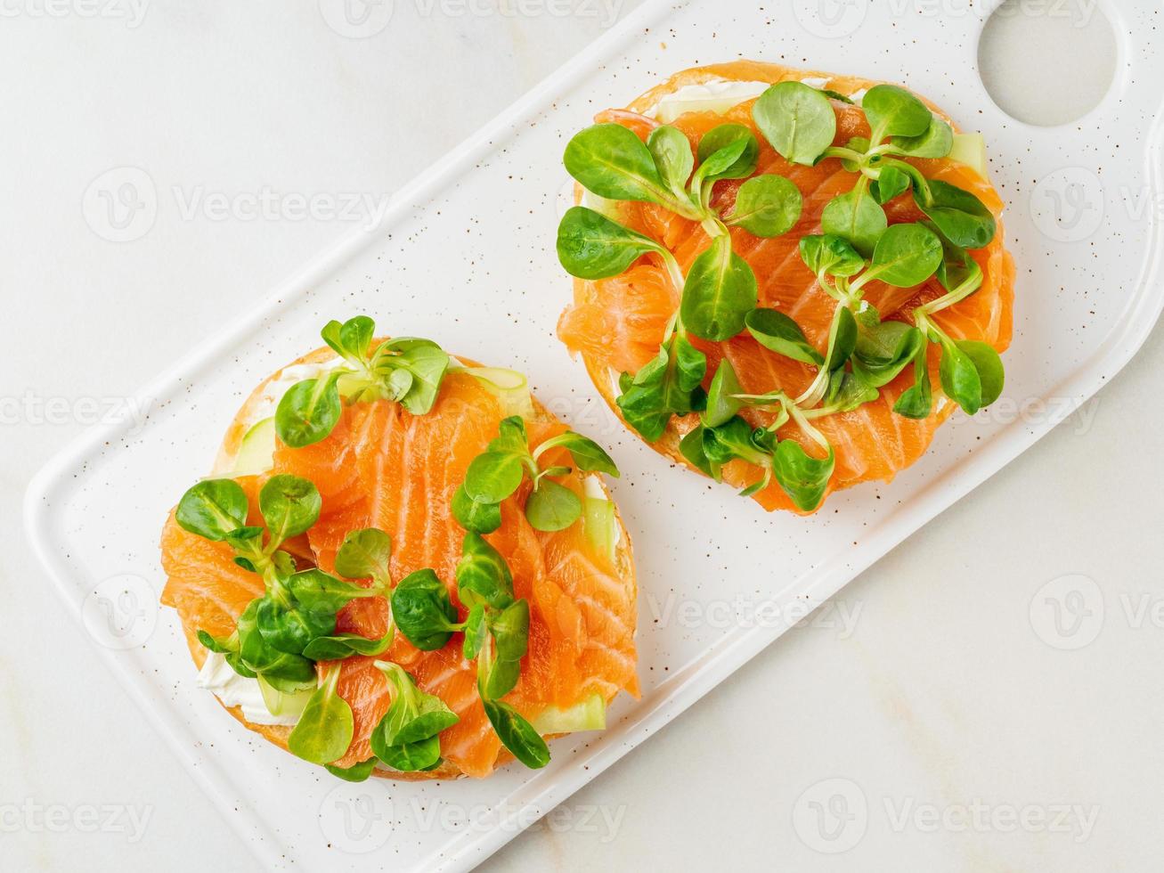 dois sanduíches abertos com salmão, cream cheese, fatias de pepino na mesa de mármore branca foto