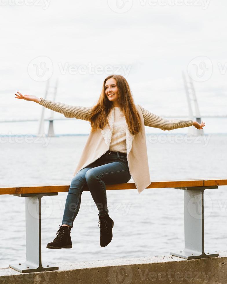 jovem feliz sentada à beira-mar no cais do porto, curtindo a vida e agitando os braços. mulher com cabelo comprido sorri e aproveita o momento foto