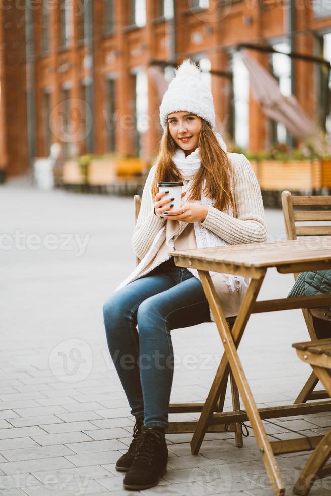 linda jovem bebendo café, chá de caneca plástica reutilizável no outono, inverno. mulher com cabelo comprido, em roupas quentes, sentado no café de rua, aquecido por bebida quente, vertical foto