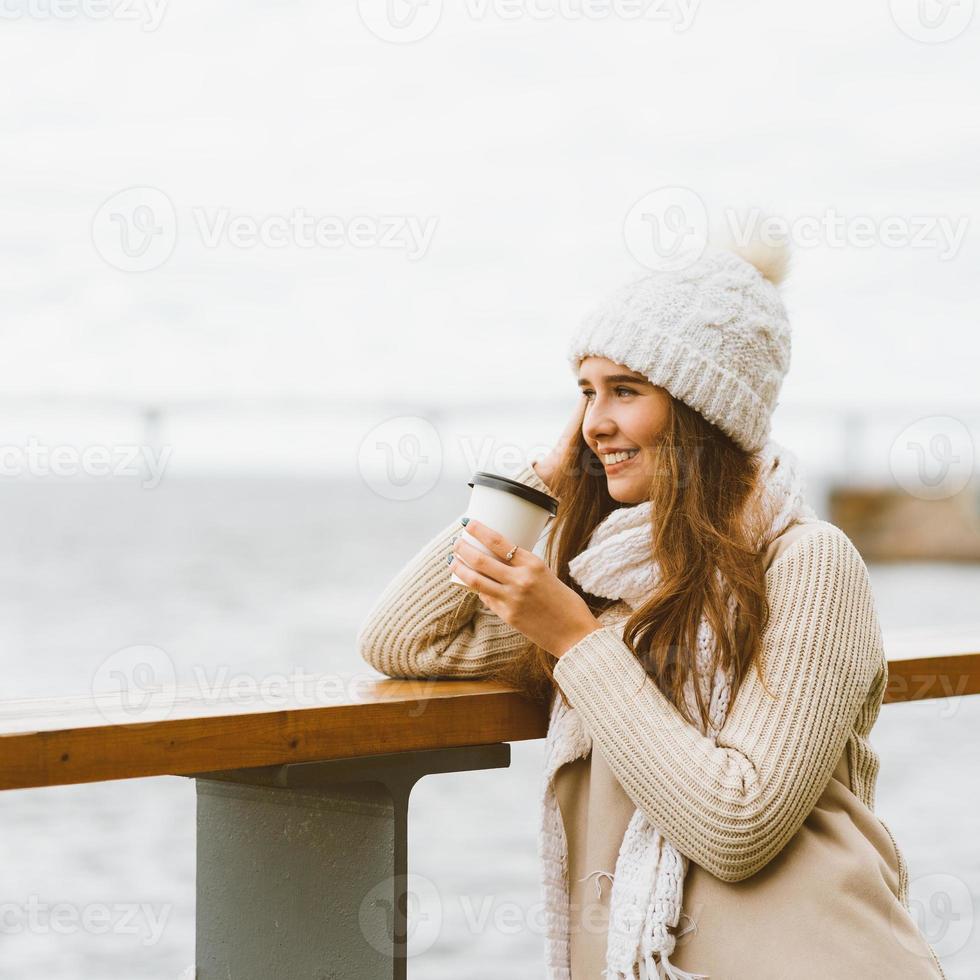 linda jovem bebendo café, chá de caneca plástica no outono, inverno. uma mulher com cabelo comprido fica à beira-mar no mar Báltico no porto e esperando a balsa, aquecida por bebida quente, copie o espaço foto