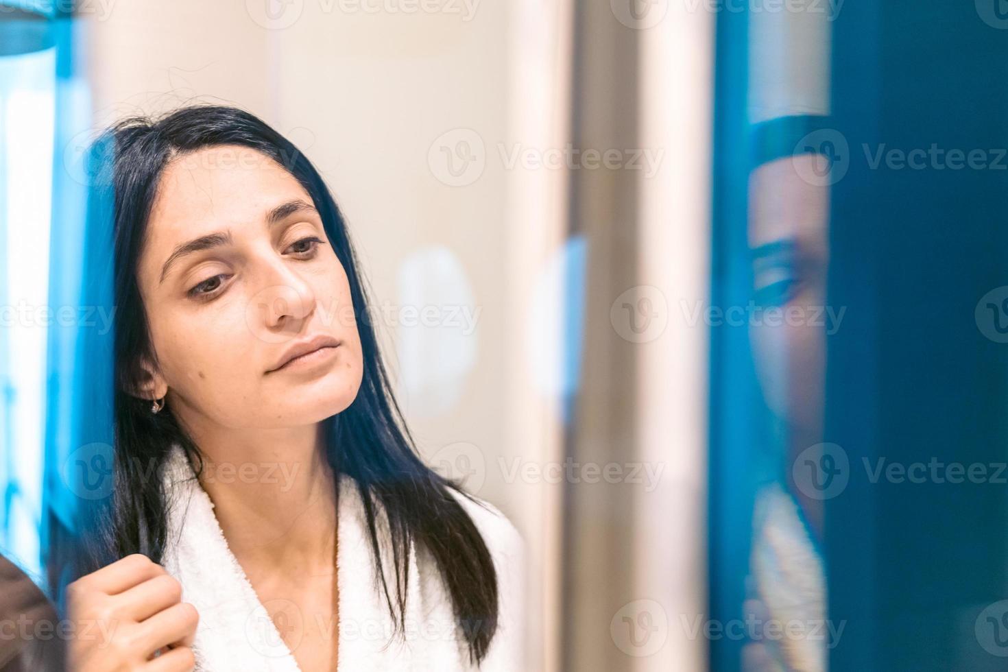 mulher bonita com rosto fresco de manhã em roupão branco olha para o espelho com desconfiança. imagem de fundo vertical foto