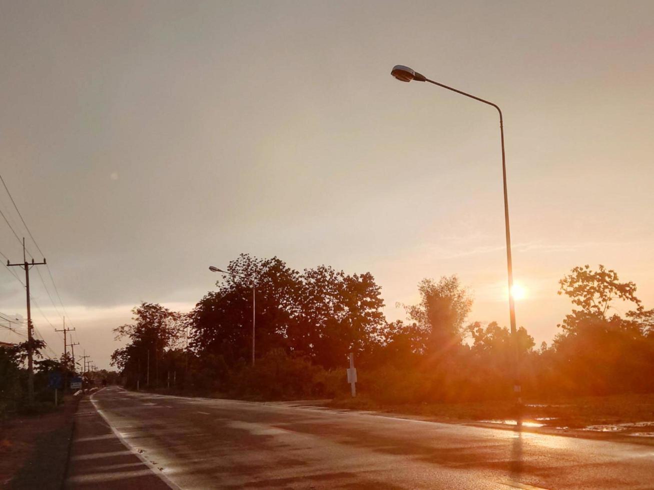 estrada pavimentada de asfalto molhada com chuva na beira da estrada ao lado da árvore do mato, silhueta pôr do sol brilha cor laranja brilhante foto