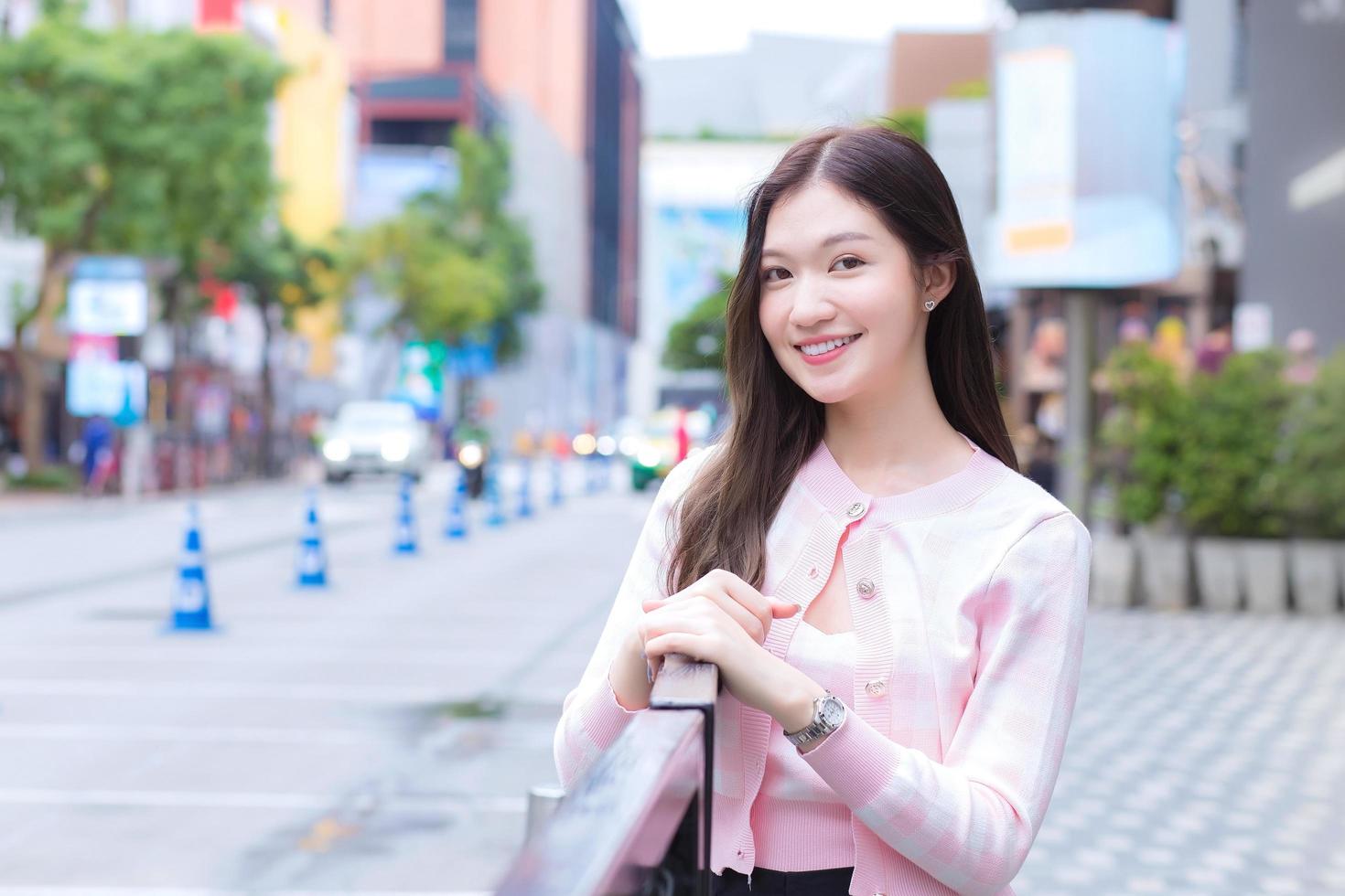 jovem mulher bonita asiática em um casaco listrado rosa e branco está de pé urbano ao ar livre ela olha para a câmera com a cidade e a estrada como pano de fundo. foto