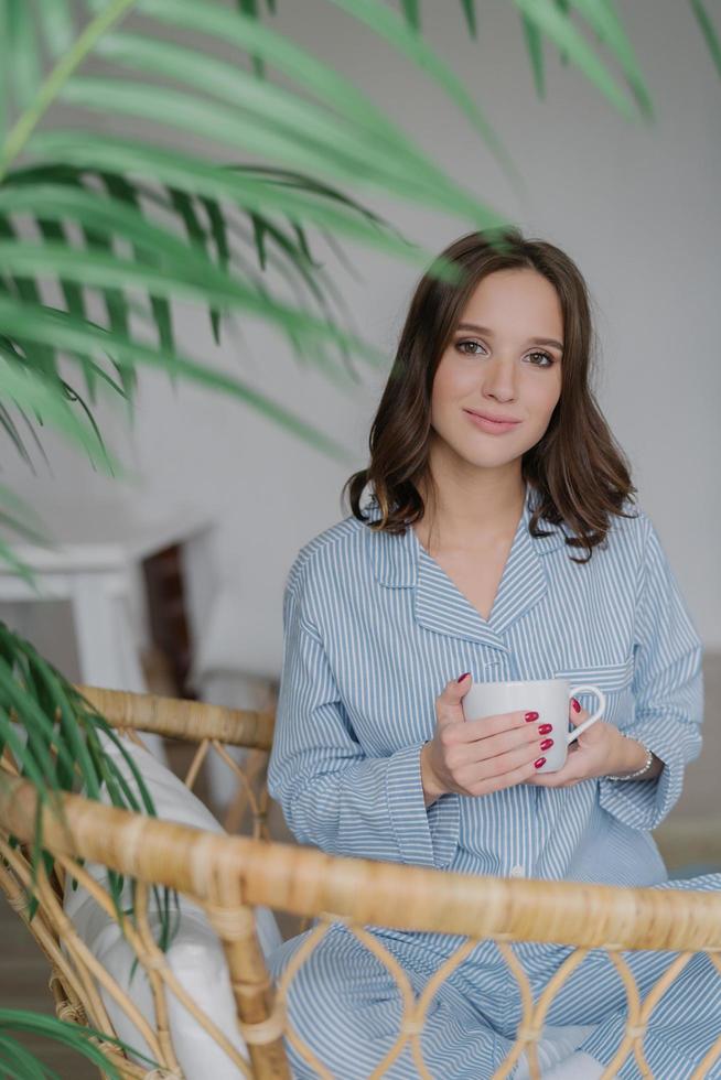 adorável jovem fêmea tem um visual encantador, vestido com traje doméstico listrado, bebe bebida quente, senta-se na cadeira de vime, gosta de descansar e dia de folga. pessoas, estilo de vida e conceito de tempo de recreação foto