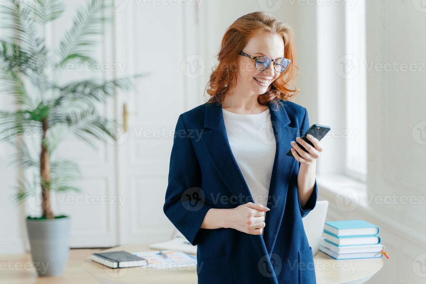 foto interna de mulher ruiva positiva verifica a caixa emial no dispositivo smartphone moderno, usa óculos ópticos, usa tecnologias modernas, recebe boas notícias, bate-papo on-line, vestido com roupas formais