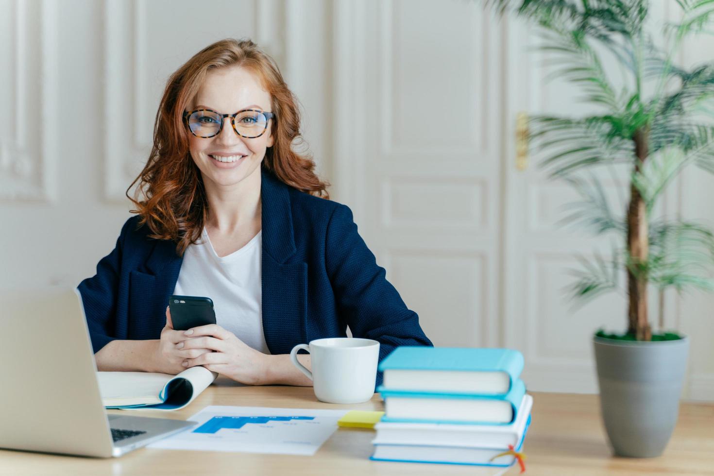 foto de jovem ruiva sorridente instala aplicativo no celular, verifica o feed de notícias, trabalha no laptop remotamente, posa no espaço de coworking com copo de bebida, livros e documentos em papel