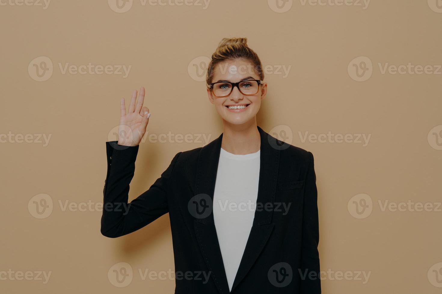 mulher caucasiana jovem positiva em desgaste clássico isolado em fundo bege, mostrando sinal de ok foto