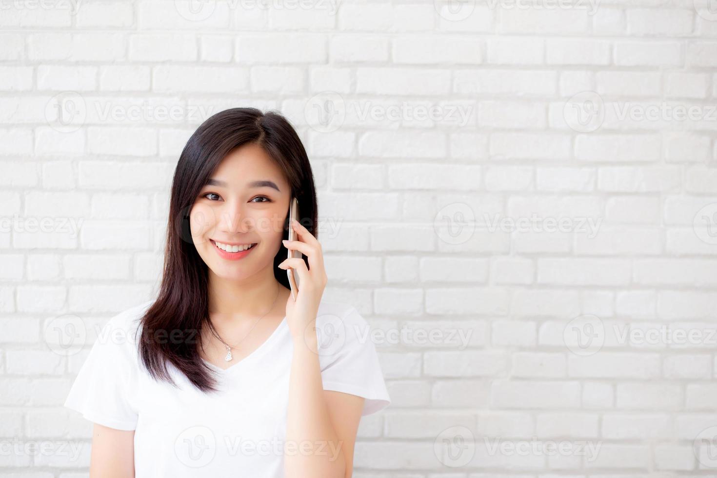 linda de retrato jovem mulher asiática falar telefone inteligente e sorrir em pé no fundo de tijolo de cimento, telefone de chamada feminina freelance, comunicação do conceito móvel. foto