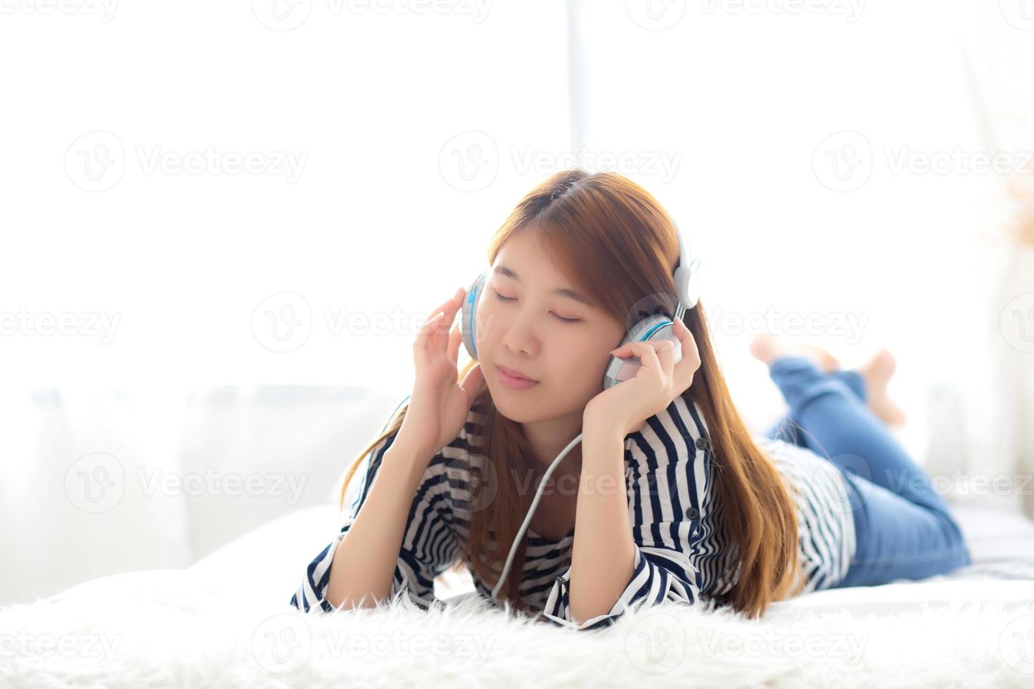 bela jovem asiática gosta e se diverte ouvindo música com fone de ouvido deitado no quarto, garota relaxa com o conceito de fone de ouvido, lazer e tecnologia. foto