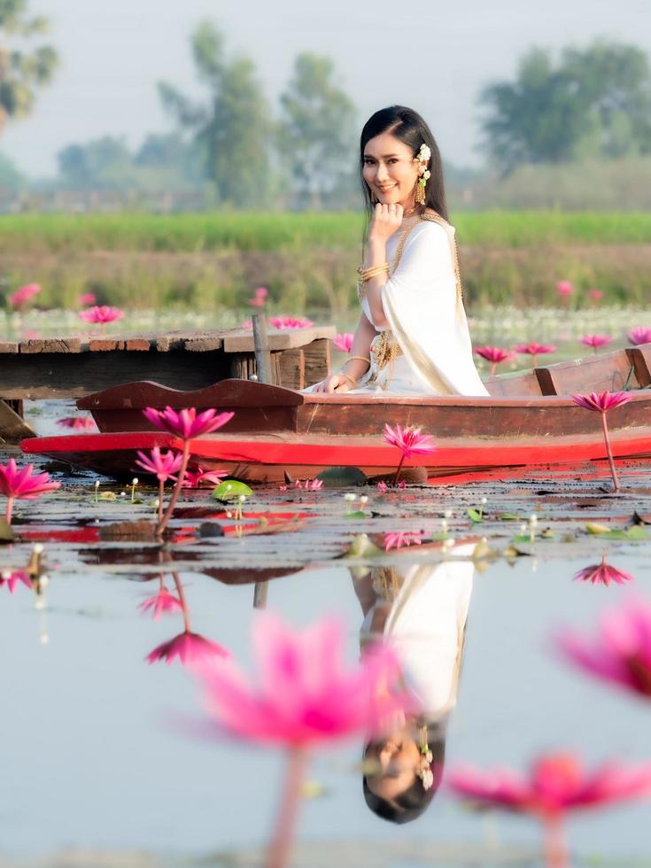 uma elegante mulher tailandesa vestindo roupas tradicionais tailandesas adornadas com ornamentos de ouro, sentada em um barco de madeira flutuando em um campo de lótus foto