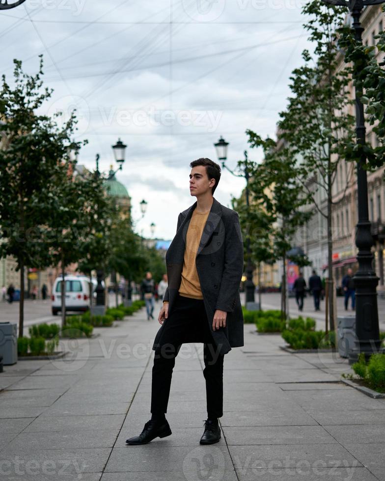homem elegante elegante bonito, morena com casaco cinza elegante, fica na rua no centro histórico de st. petersburgo. jovem com cabelos escuros, sobrancelhas grossas. foto