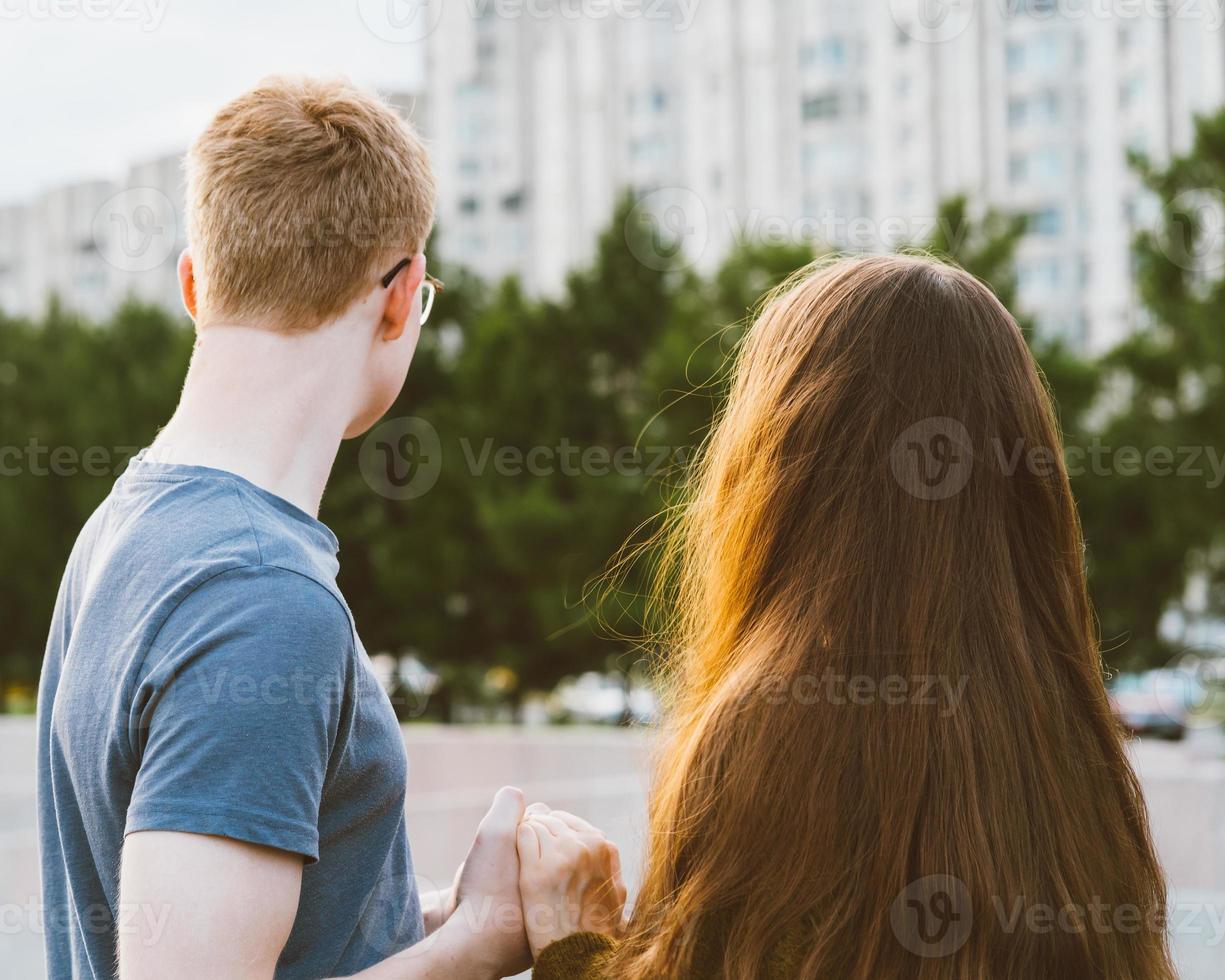 menina com cabelo escuro longo e grosso, de mãos dadas, menino ruivo de camiseta azul na ponte, amor adolescente à noite. menino olha com ternura para a menina, jovem casal. conceito de amor adolescente e primeiro beijo foto