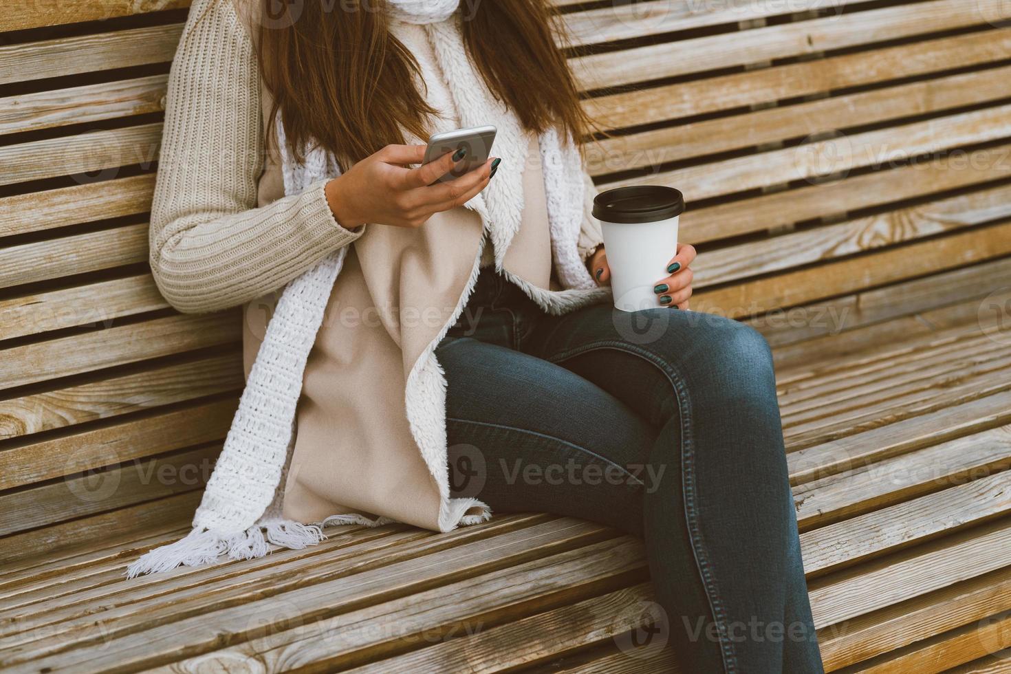 linda jovem irreconhecível bebendo café, chá de caneca de plástico no outono, inverno e falando no celular. mulher com cabelo comprido sentado no banco no outono ou inverno, tomando uma bebida quente foto