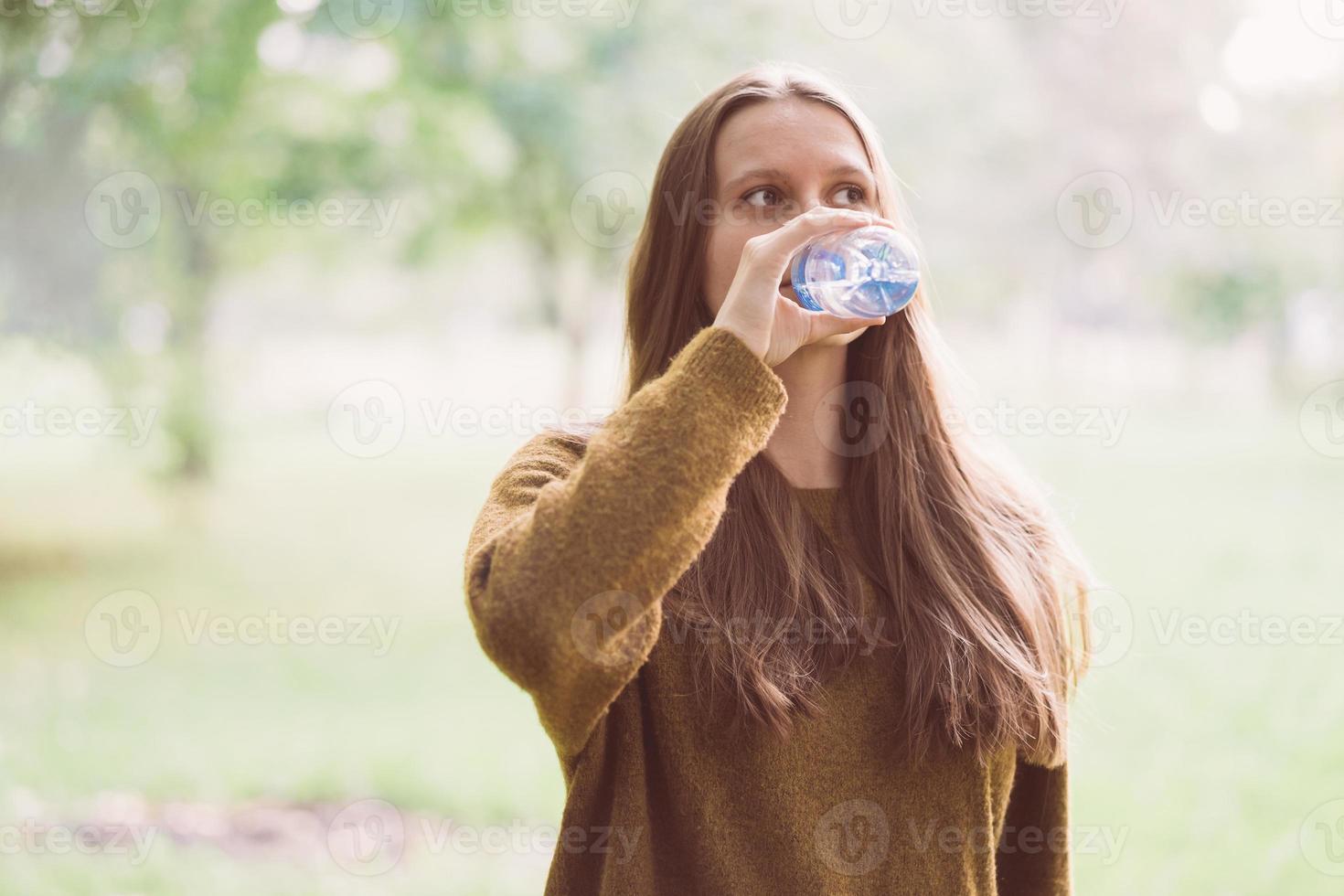 linda jovem bebendo água de uma garrafa de plástico na rua no parque no outono ou inverno. uma mulher com lindos cabelos escuros longos e grossos sacia sua sede de água em uma caminhada foto