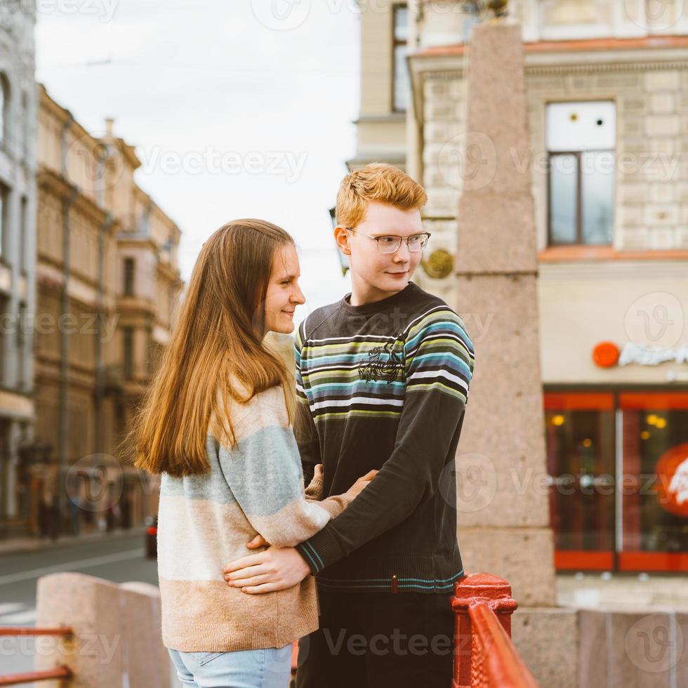menina com muito espesso escuro ouvir abraçando menino ruivo de camiseta azul na ponte, jovem casal. conceito de amor adolescente. eles desviam o olhar, viram alguma coisa. cidade, beira-mar. foto