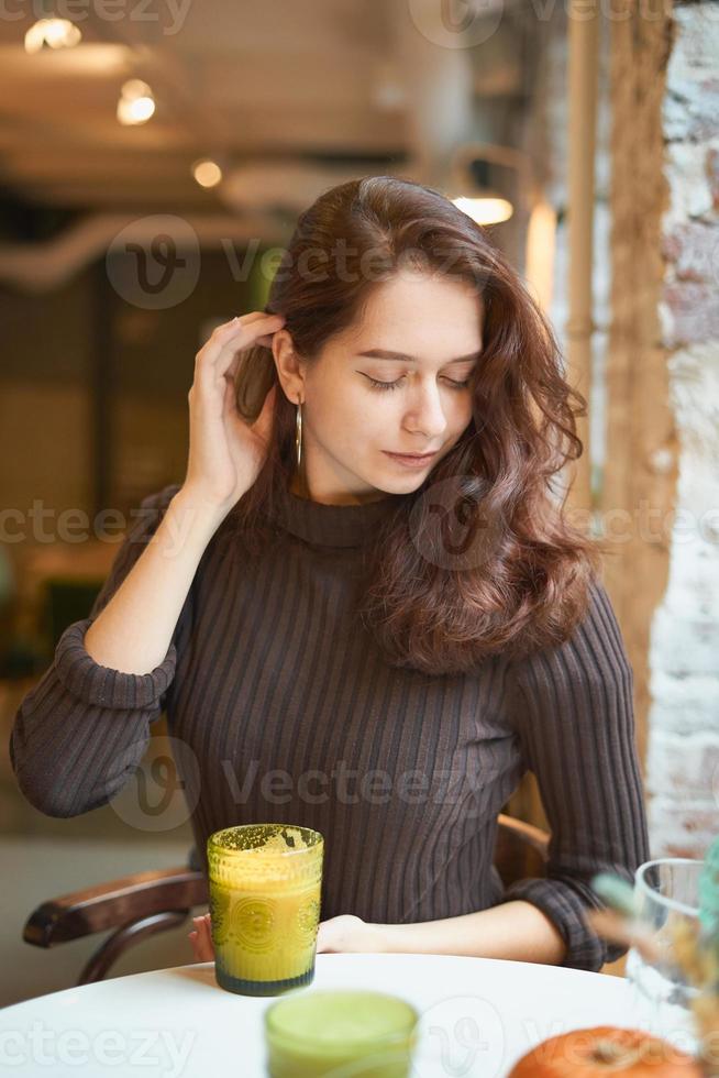 linda garota inteligente elegante elegante está sentada no café e bebendo smoothie amarelo saudável ou latte vegan. encantadora mulher pensativa com longos cabelos castanhos escuros. foto