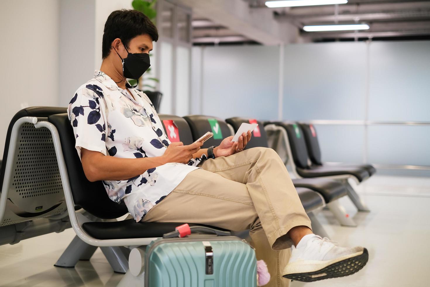 viajante asiático usando máscara protetora usando smartphone pesquisando o status do voo da companhia aérea e sentado com distância no aeroporto internacional. novo normal e distanciamento social. foto