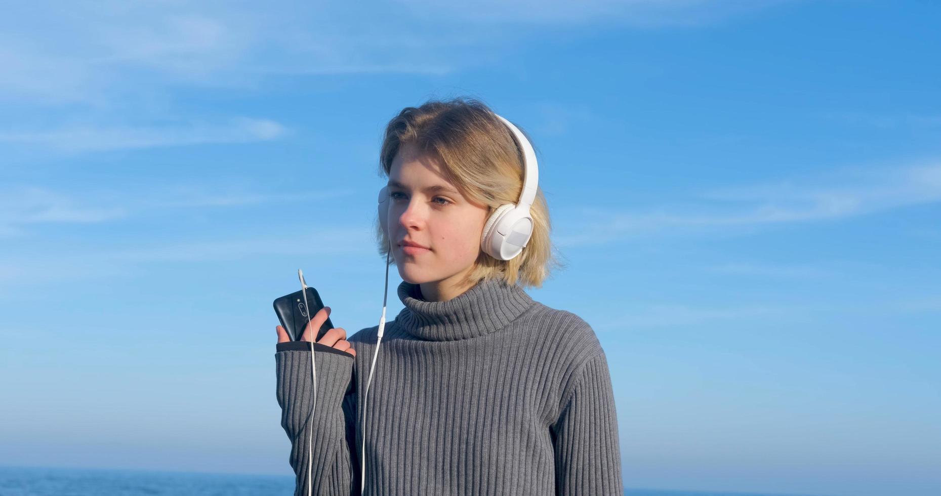 jovem fêmea bonita ouvir música com fones de ouvido ao ar livre na praia contra o céu azul ensolarado foto