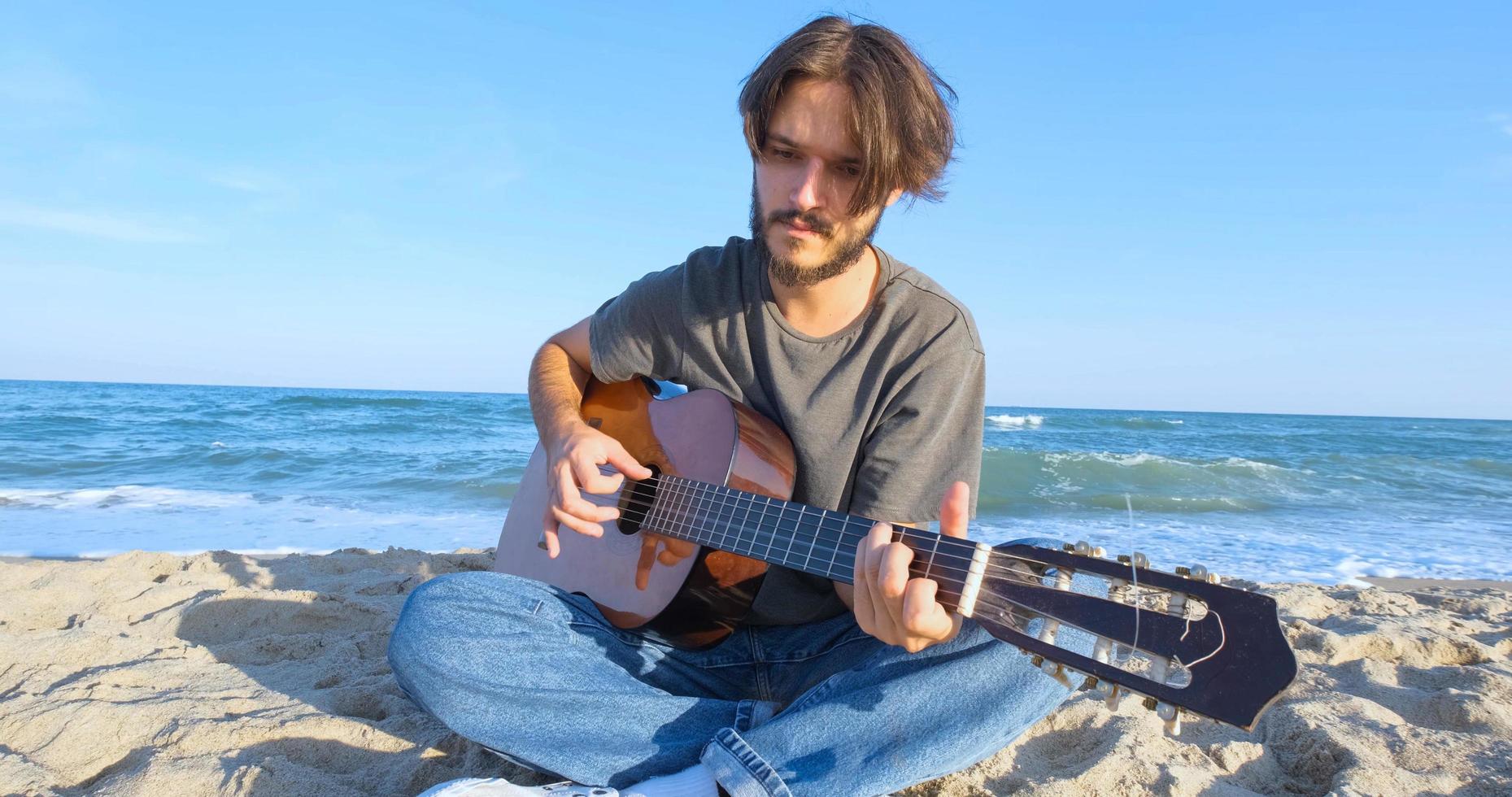 jovem macho bonito toca violão na praia em dia ensolarado, mar ou oceano no fundo foto