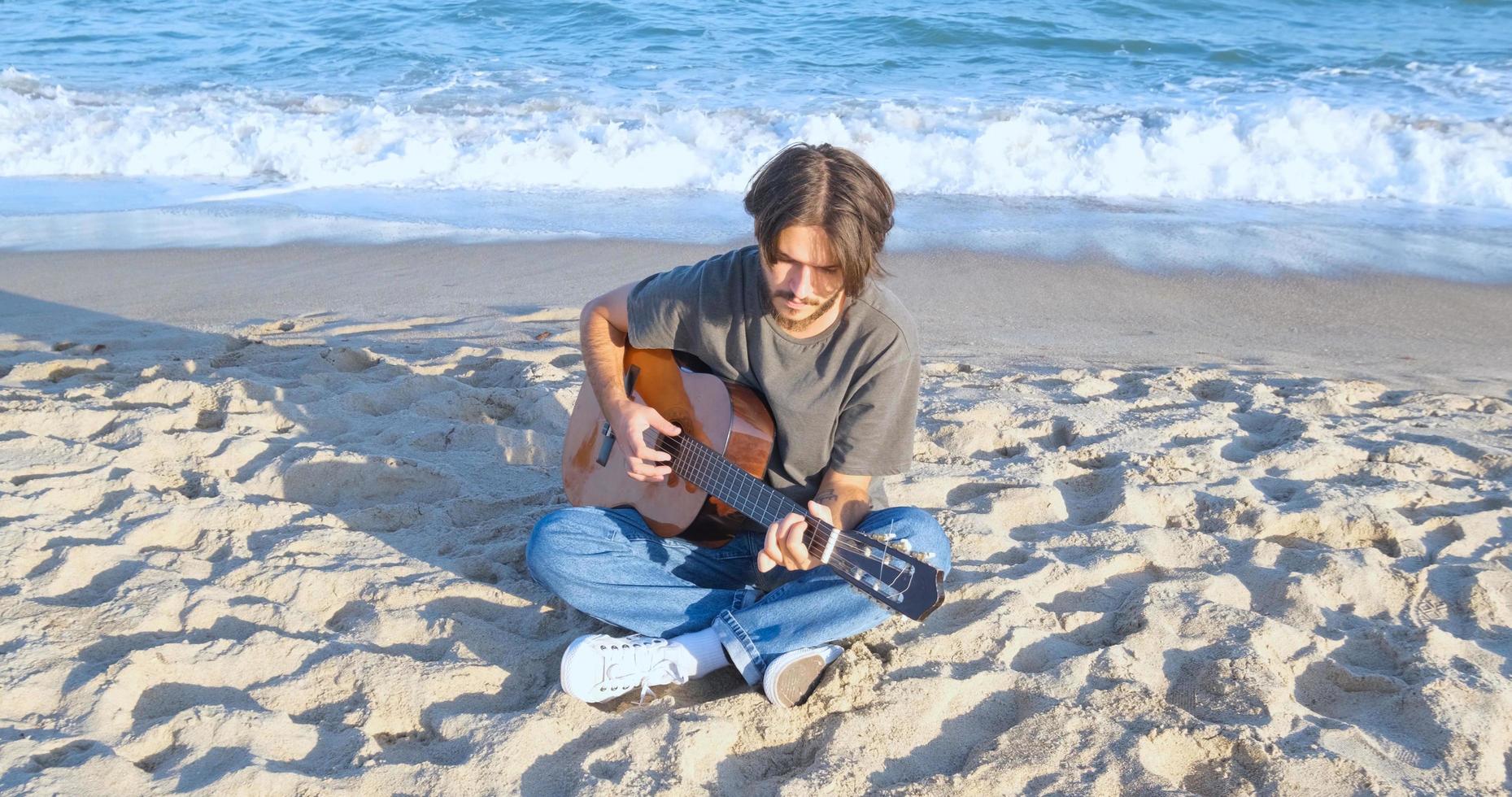 jovem macho bonito toca violão na praia em dia ensolarado, mar ou oceano no fundo foto