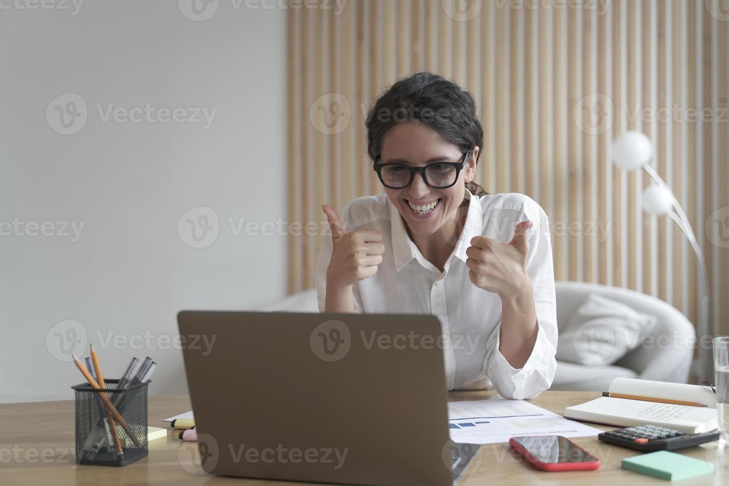 mulher de negócios olhando para a tela do computador com um sorriso largo enquanto mostra o polegar para cima com as duas mãos foto