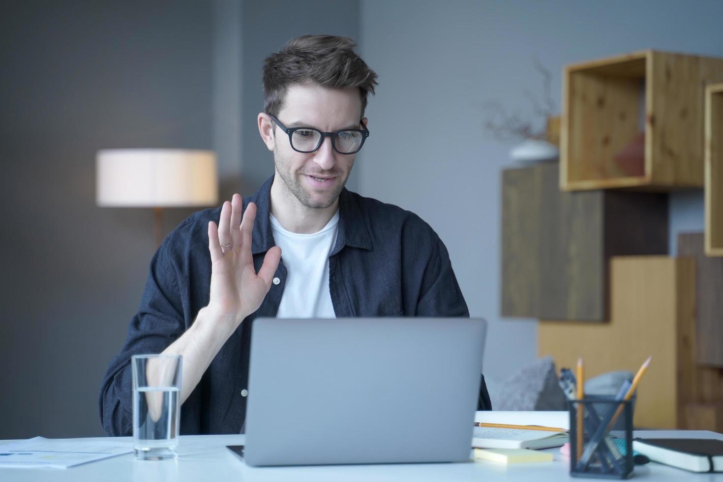 jovem austríaco bonito freelancer acenando com a mão no gesto de olá durante a chamada de vídeo no laptop foto