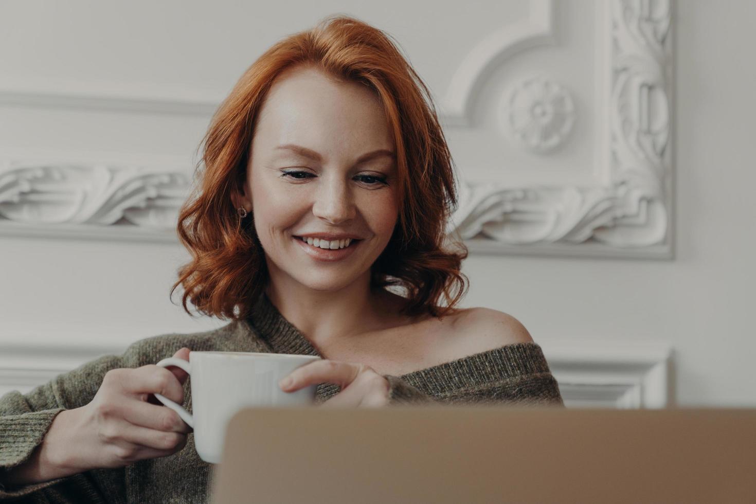 tiro horizontal de mulher bonita ruiva assiste a webinar de treinamento no computador portátil, bebe bebidas refrescantes, procura informações para o projeto, trabalha em casa, compartilha fotos em redes sociais