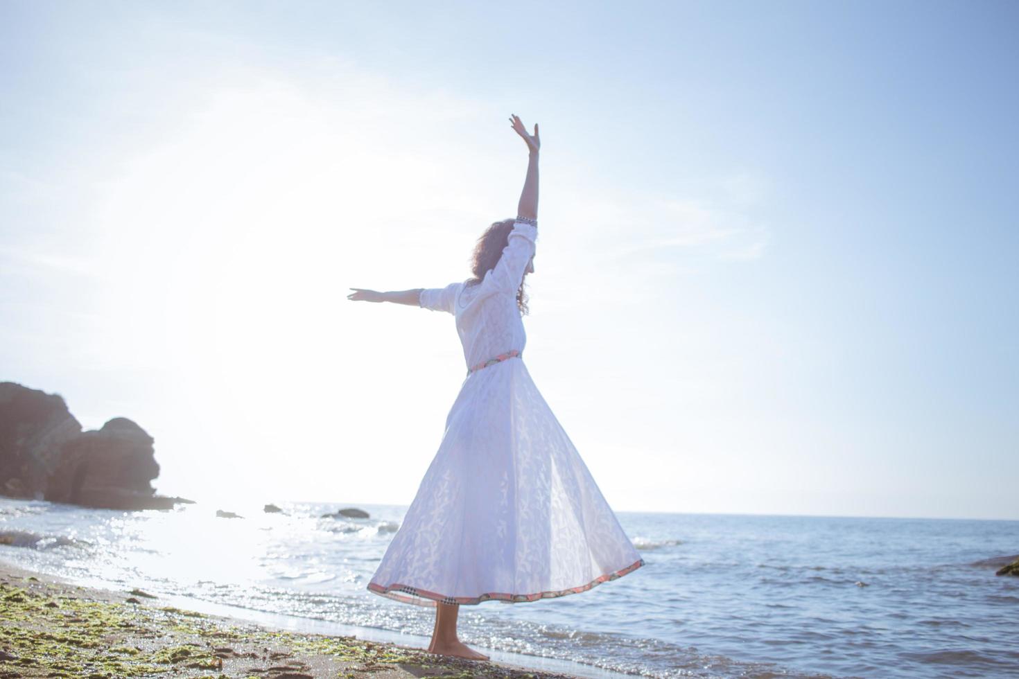 jovem caminhando na praia de manhã em lindo vestido branco. apto feminino se divertindo durante o nascer do sol. foto