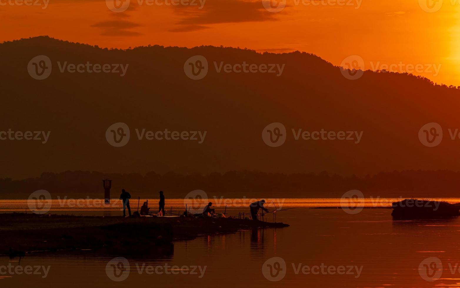 lindo céu do nascer do sol acima da montanha no reservatório. as pessoas estão pescando com uma vara de pescar no rio. paisagem de reservatório e montanha com céu laranja do nascer do sol. vida de silhueta pela manhã. foto