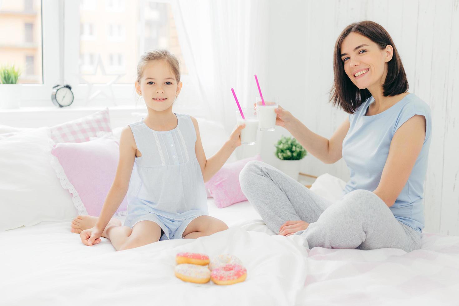 retrato de mãe jovem feliz e sua filha pequena seguram copos de milk-shake, tomam café da manhã na cama, olham positivamente para a câmera, aproveita o fim de semana em casa e união, vestido com roupa de dormir foto