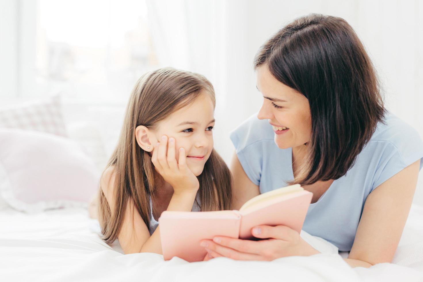 A filhinha satisfeita olha curiosamente para a mãe que lê conto de fadas, segura um pequeno livro, deita na cama confortável, desfruta da união. criança alegre se prepara para a escola. conceito de paternidade foto