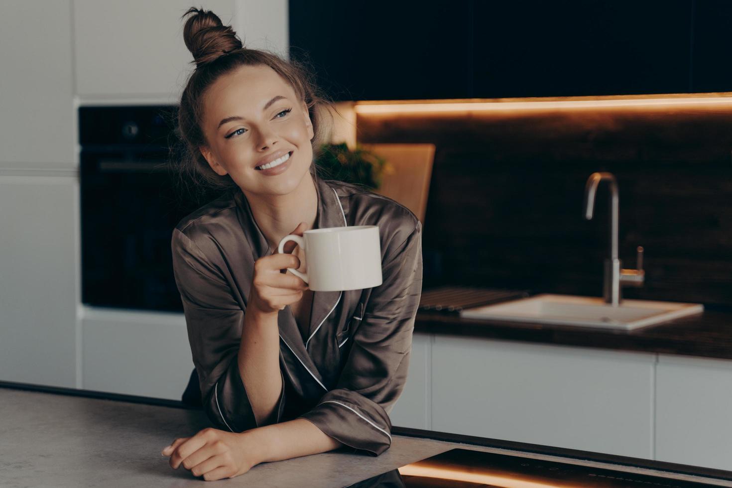 feliz sorridente jovem de pijama de cetim marrom, desfrutando de café aromático da manhã na cozinha foto