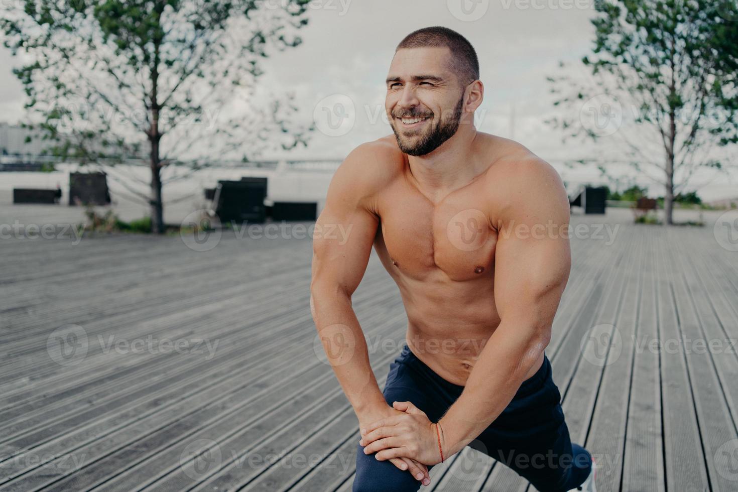 rotina matinal diária e conceito de exercícios esportivos. fisiculturista de homem com barba esportiva com expressão alegre faz exercícios de alongamento para pernas, poses com torso nu, treino ao ar livre. foto