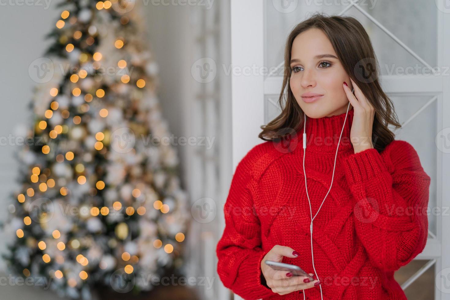 pessoas, feriados e conceito de tecnologia. mulher bonita usa telefone celular e fones de ouvido para ouvir música, fica em casa contra o fundo das luzes da árvore de natal com espaço de cópia para o seu texto foto