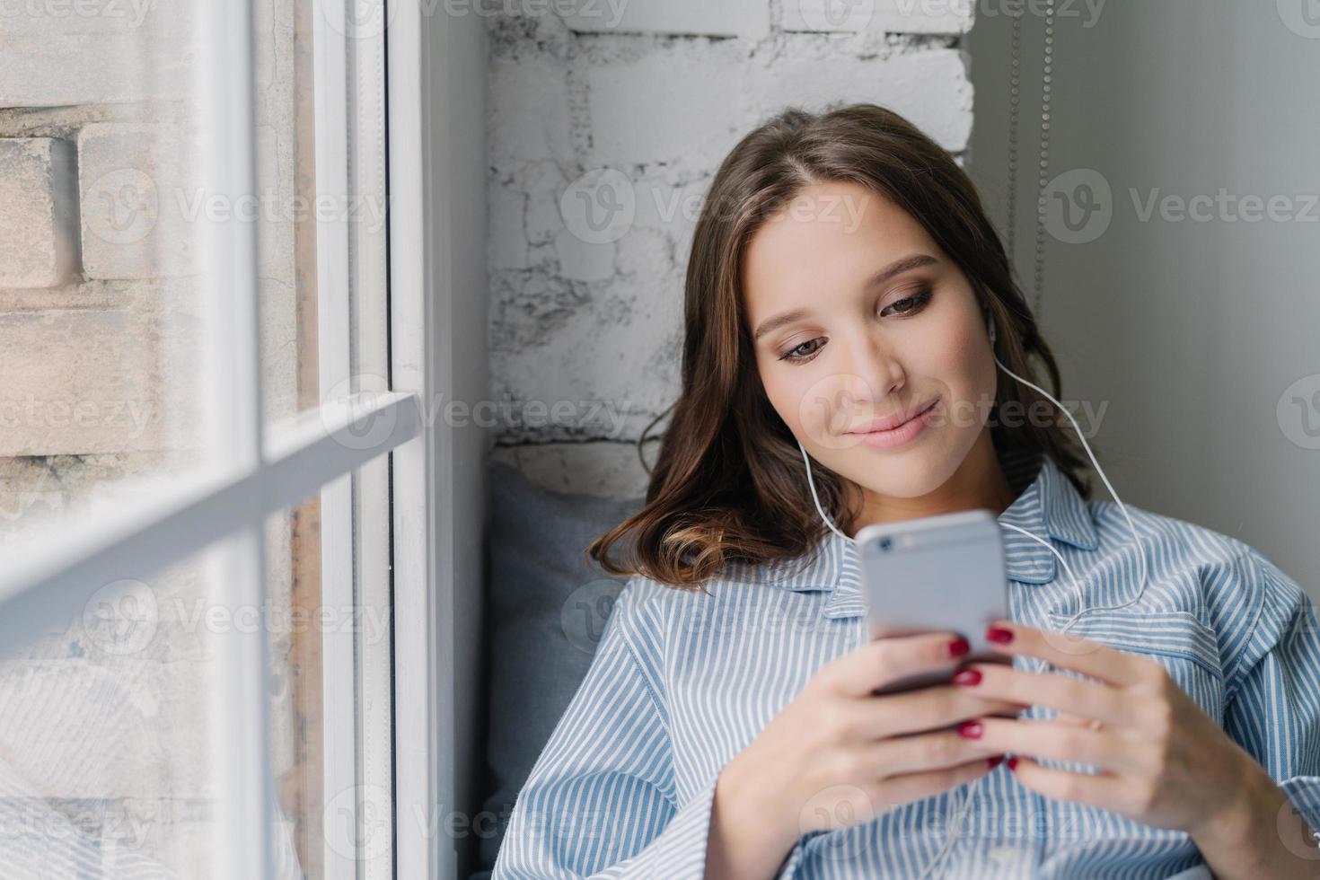 mulher atraente sorridente cria nova lista de reprodução no celular, digita e-mail e gosta de música em fones de ouvido, senta-se no peitoril da janela em um quarto aconchegante, gosta de lonilenss. linda garota faz videochamada em casa foto