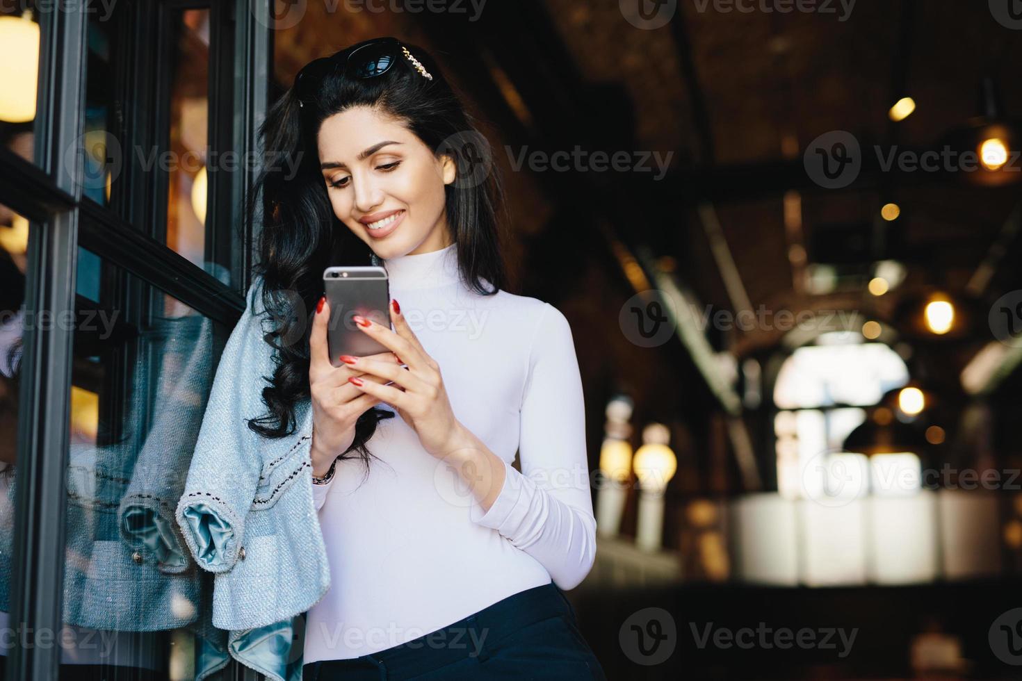 adorável mulher morena com olhar feliz com óculos escuros na cabeça e blusa branca localização sobre o interior do café moderno, usando telefone celular, verificando o feed de notícias em suas contas de redes sociais. foto