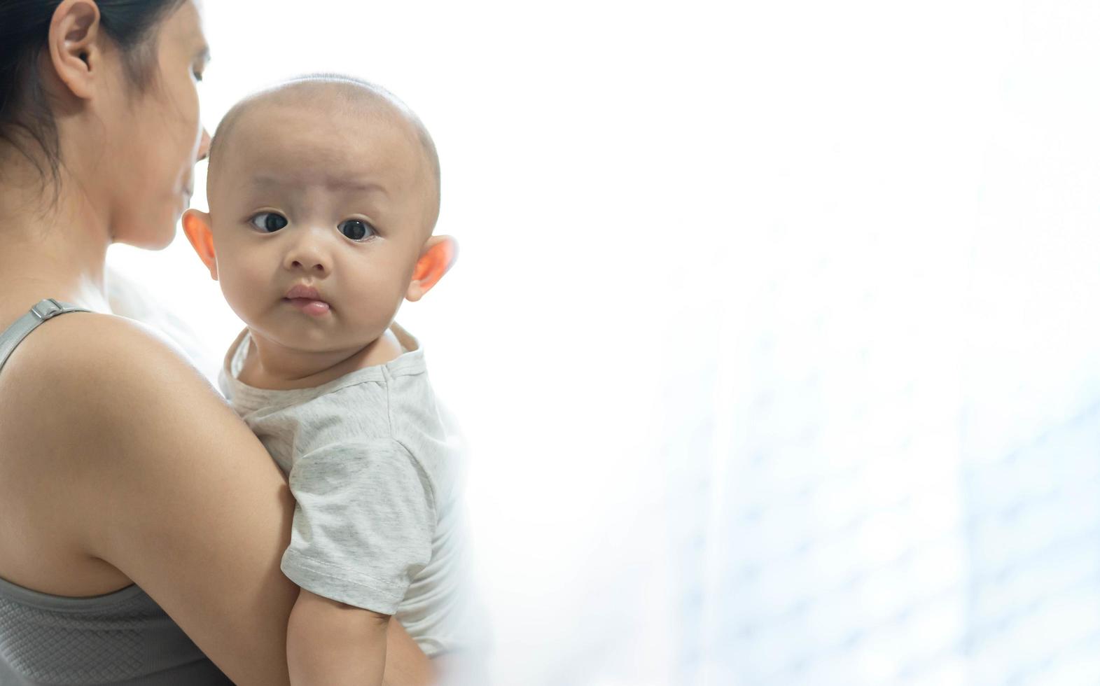 menino asiático bonito e recém-nascido. família feliz, jovem mãe asiática em pé carrega criança perto da janela, carinha feliz. pequena criança inocente no primeiro dia de vida. conceito de dia das mães. foto