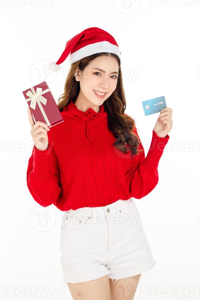 uma linda mulher asiática usando um vestido vermelho com um chapéu de papai noel está segurando uma caixa de presente. conceito de compras. foto