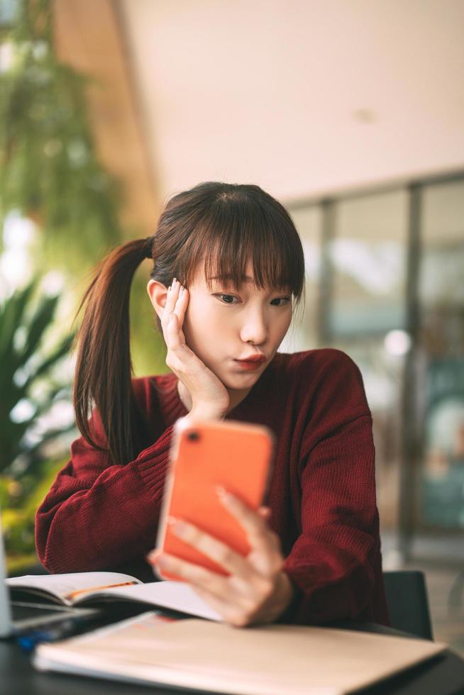 jovem estudante asiática adulta usa suéter vermelho usando telefone celular e laptop para estudar on-line no café foto
