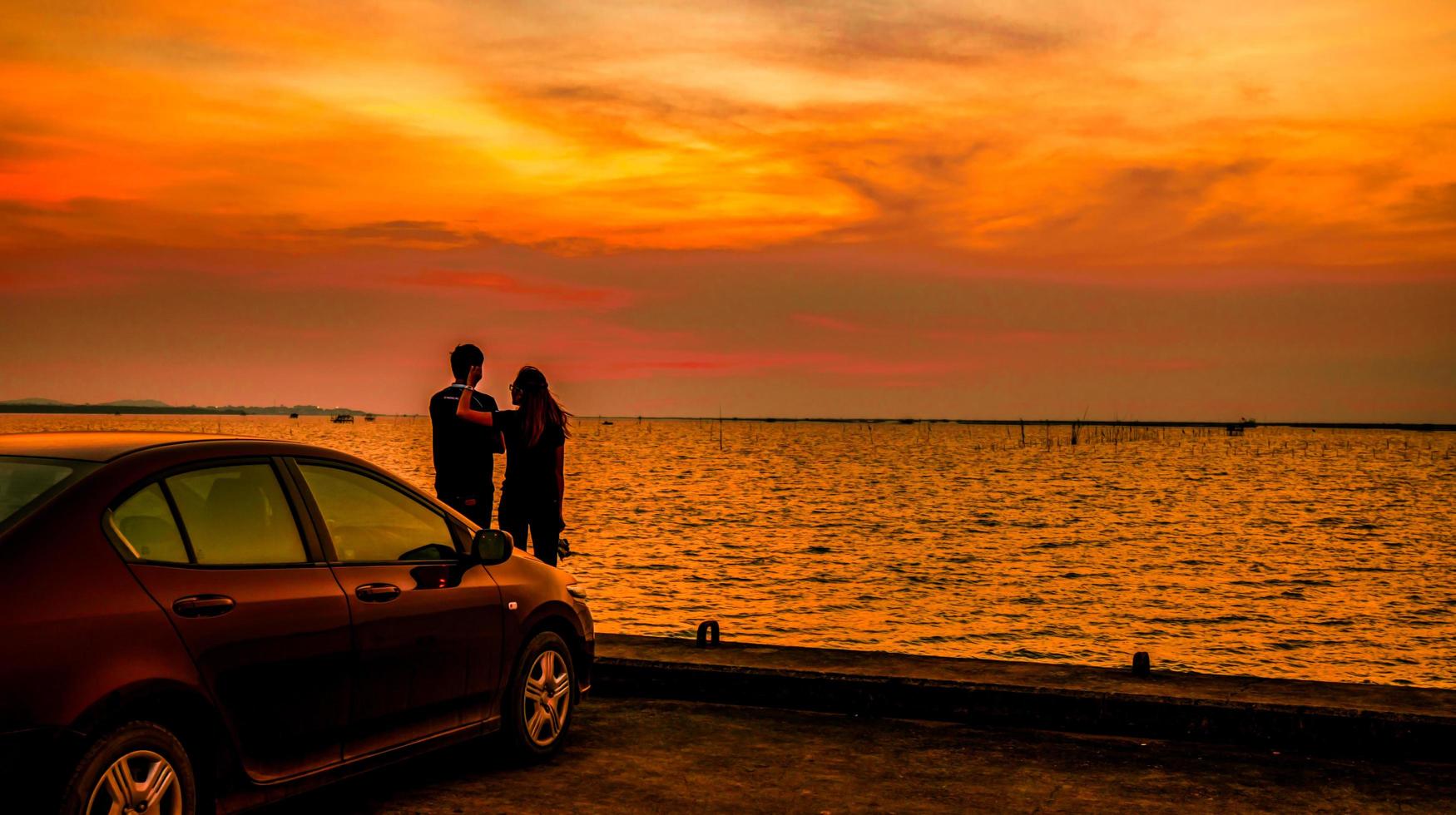 silhueta de casal de felicidade ao lado do carro à beira-mar ao pôr do sol. lindo céu laranja e nuvens foto