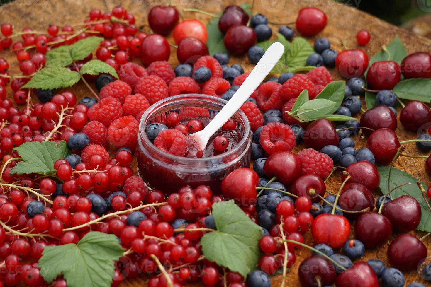 colher branca com geléia de frutas no copo. mirtilo, framboesa, cereja e groselha na mesa de madeira no jardim. foto